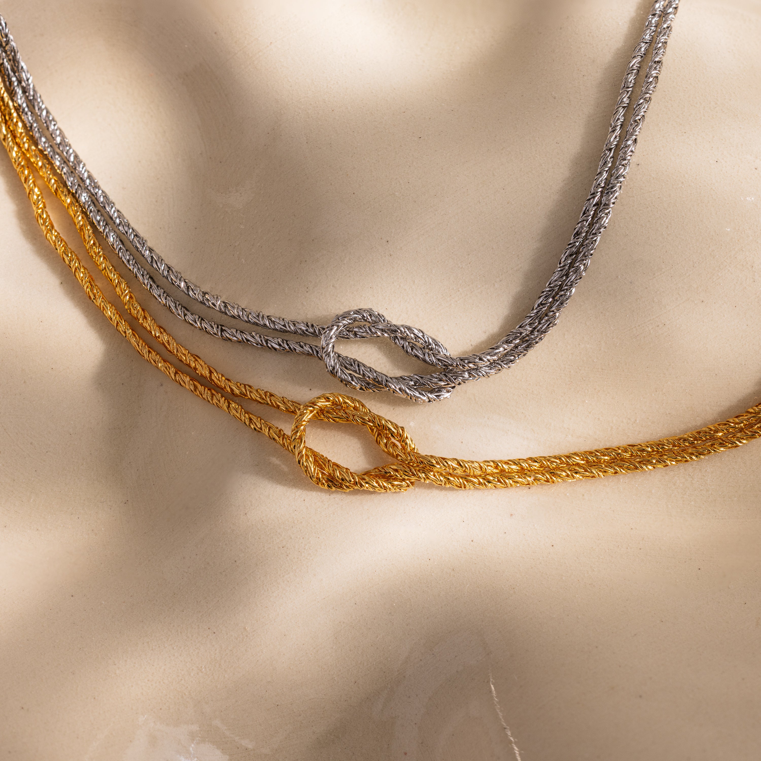 Close-up of two Rope Knot Chain Bracelets in gold and silver, each with a center knot detail, displayed on a neutral background.