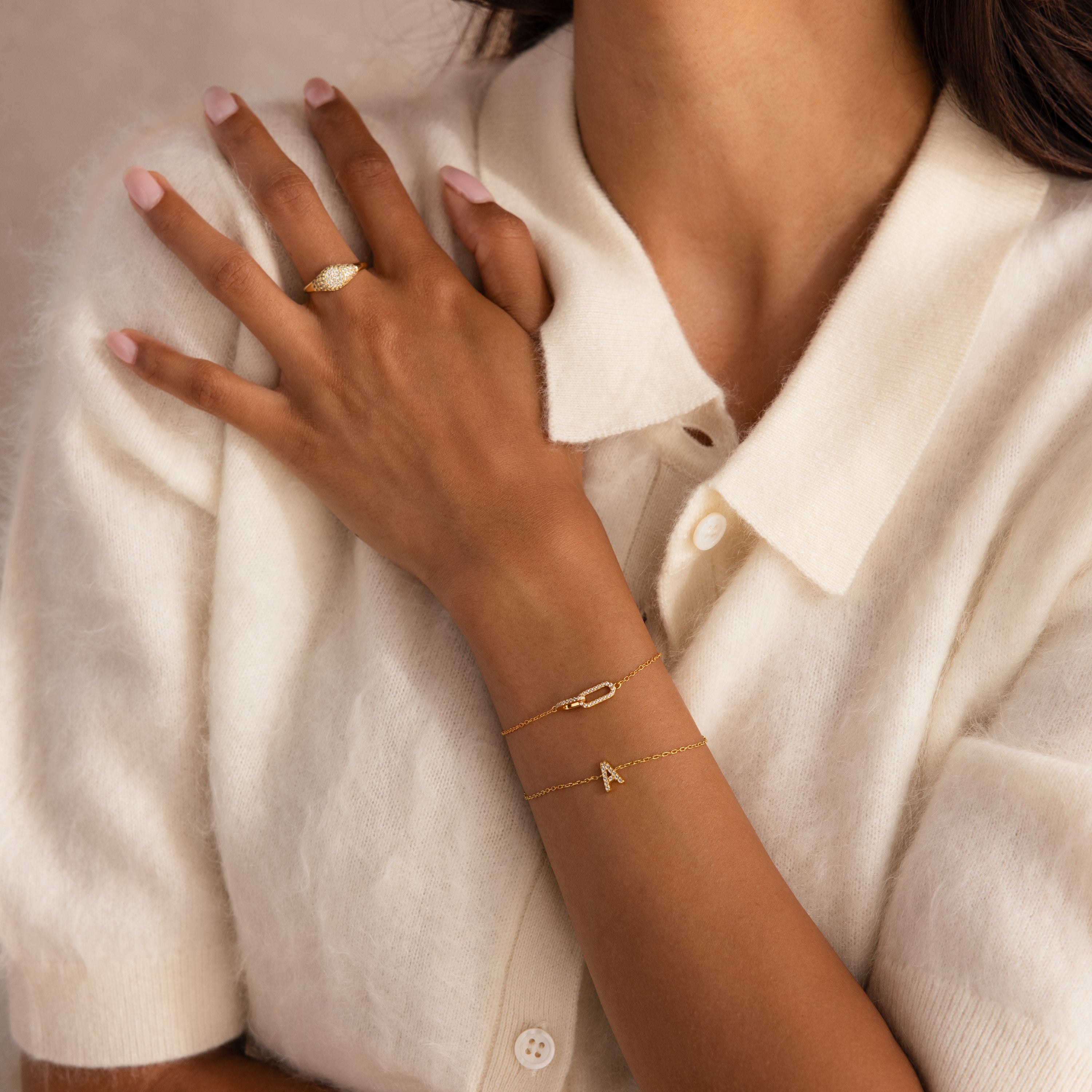 Model wearing a gold chain bracelet featuring two interlocking pave links charms, styled with a pave initial 'A' bracelet and pave signet ring.