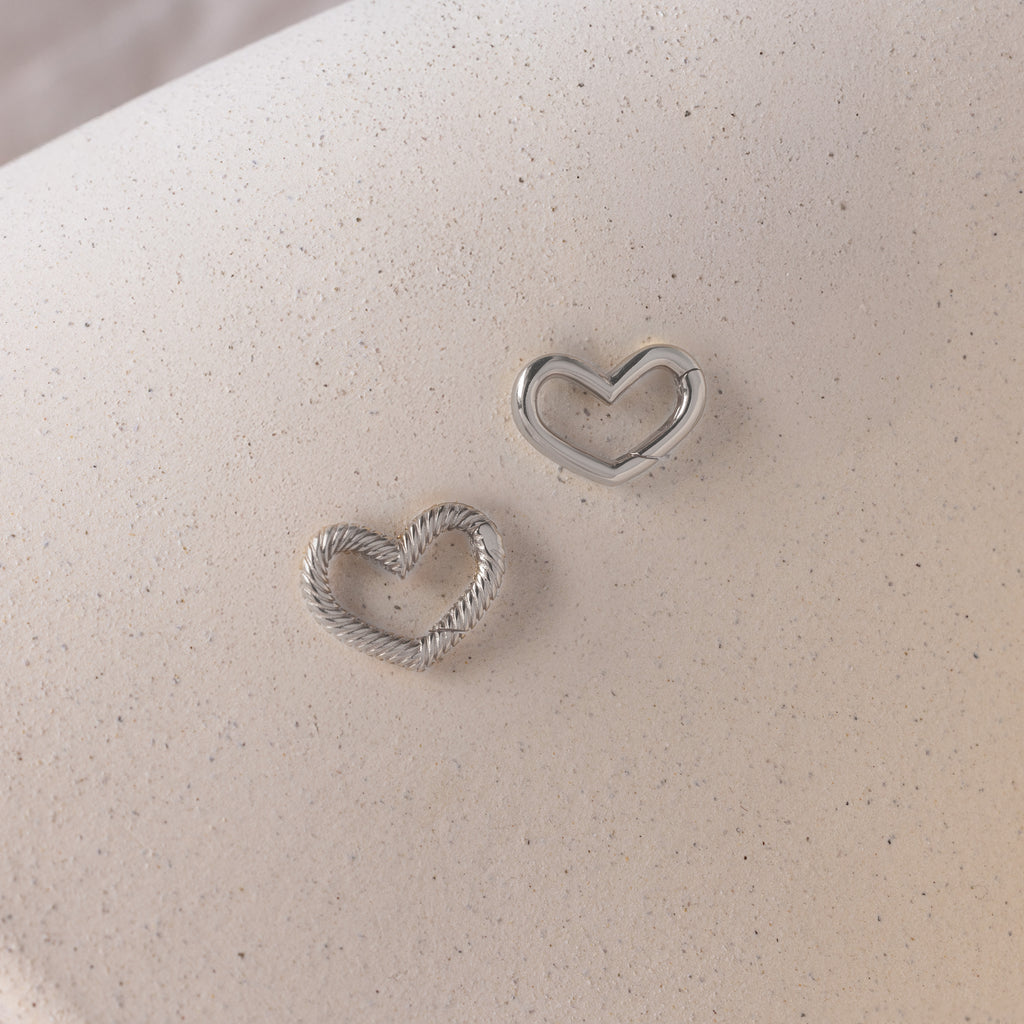 Two silver heart-shaped earrings rest on a light beige, textured surface—ideal for pairing with the Heart Carabiner Set in Sterling Silver or enhancing your collection of heart-themed jewelry.