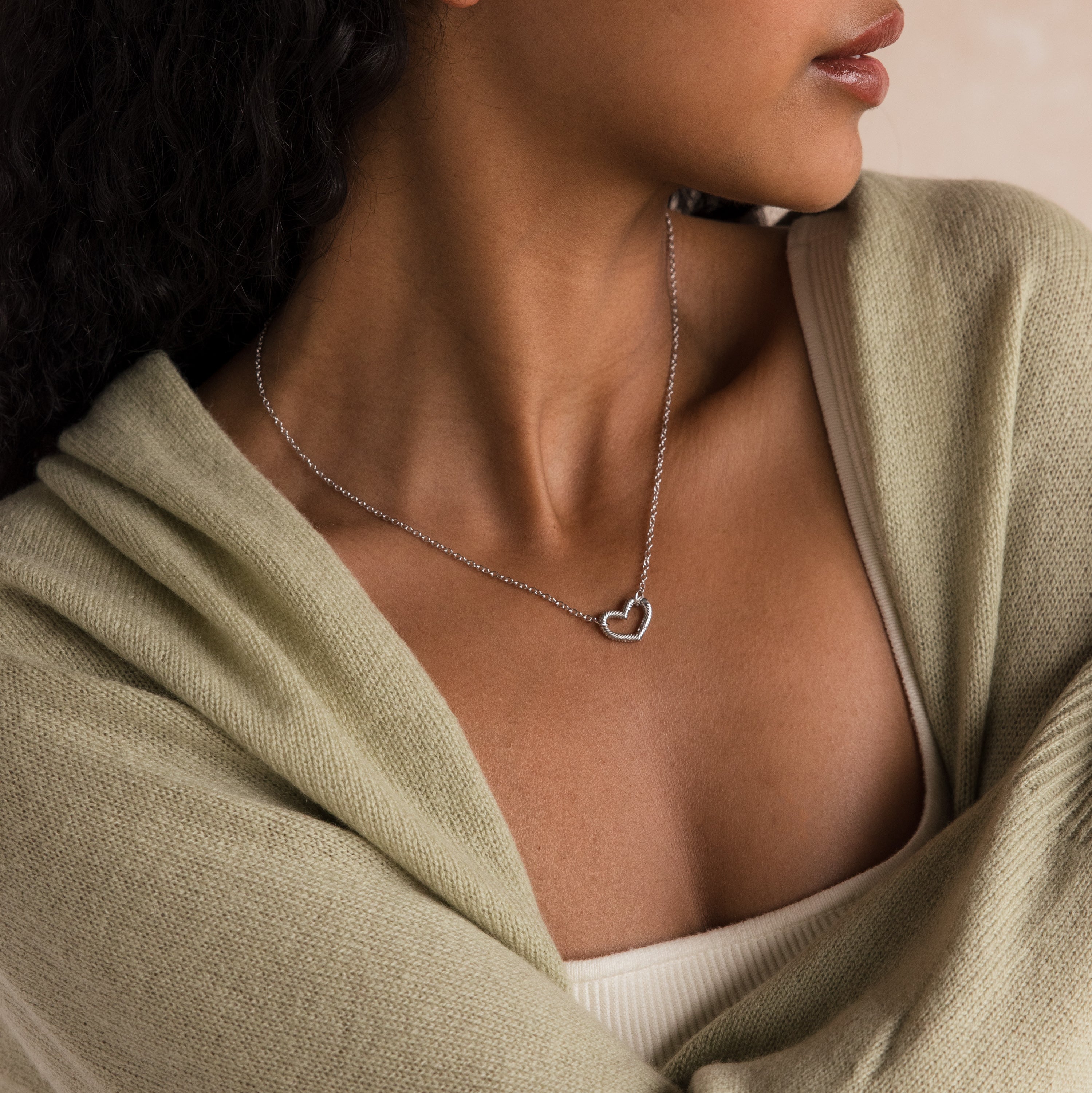 Woman wears the Heart Carabiner Set in Sterling Silver necklace, paired with a light green shawl and cream top, as she gazes to the side.