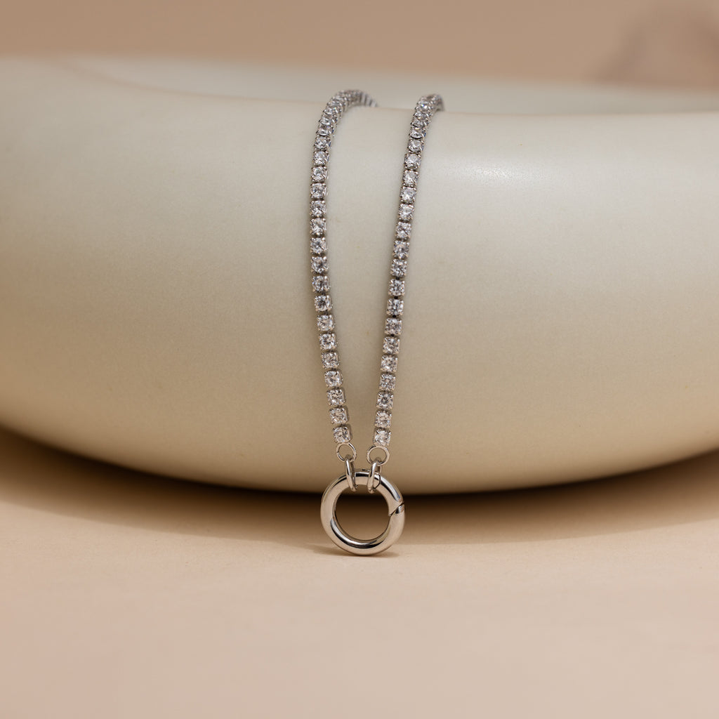 The Diamond Tennis Clasp Necklace in White Gold is draped over a smooth, light beige ceramic surface.