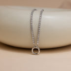 The Diamond Tennis Clasp Necklace in White Gold is draped over a smooth, light beige ceramic surface.