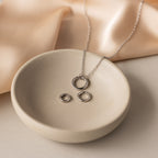 The Round Carabiner Charm Set in Sterling Silver, featuring a necklace and two matching rings, rests in a ceramic dish against a soft beige fabric backdrop.