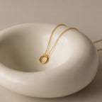 The Round Carabiner Charm Set in 18K Gold, featuring a circular pendant, rests on a smooth white ceramic dish.