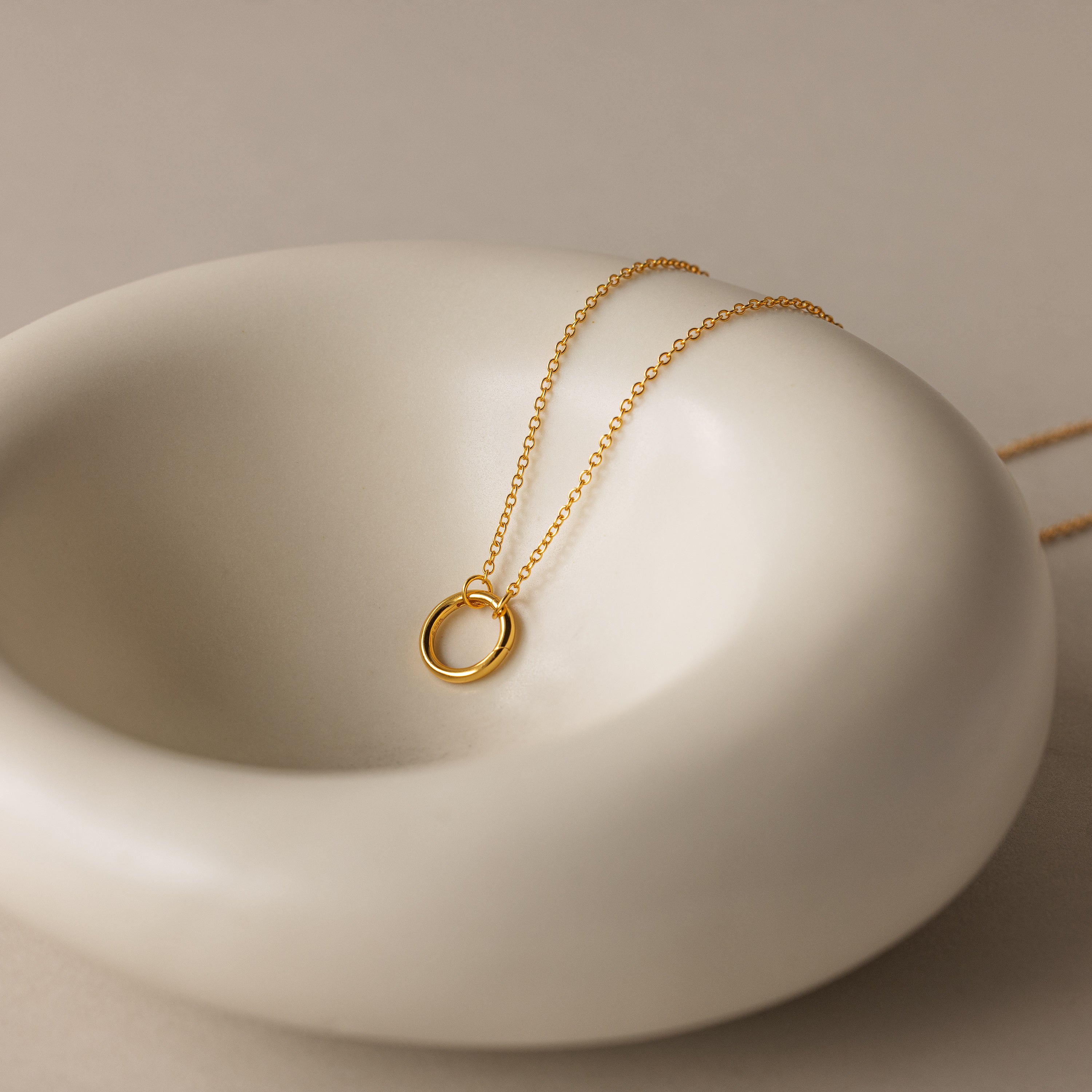 The Round Carabiner Charm Set in 18K Gold, featuring a circular pendant, rests on a smooth white ceramic dish.