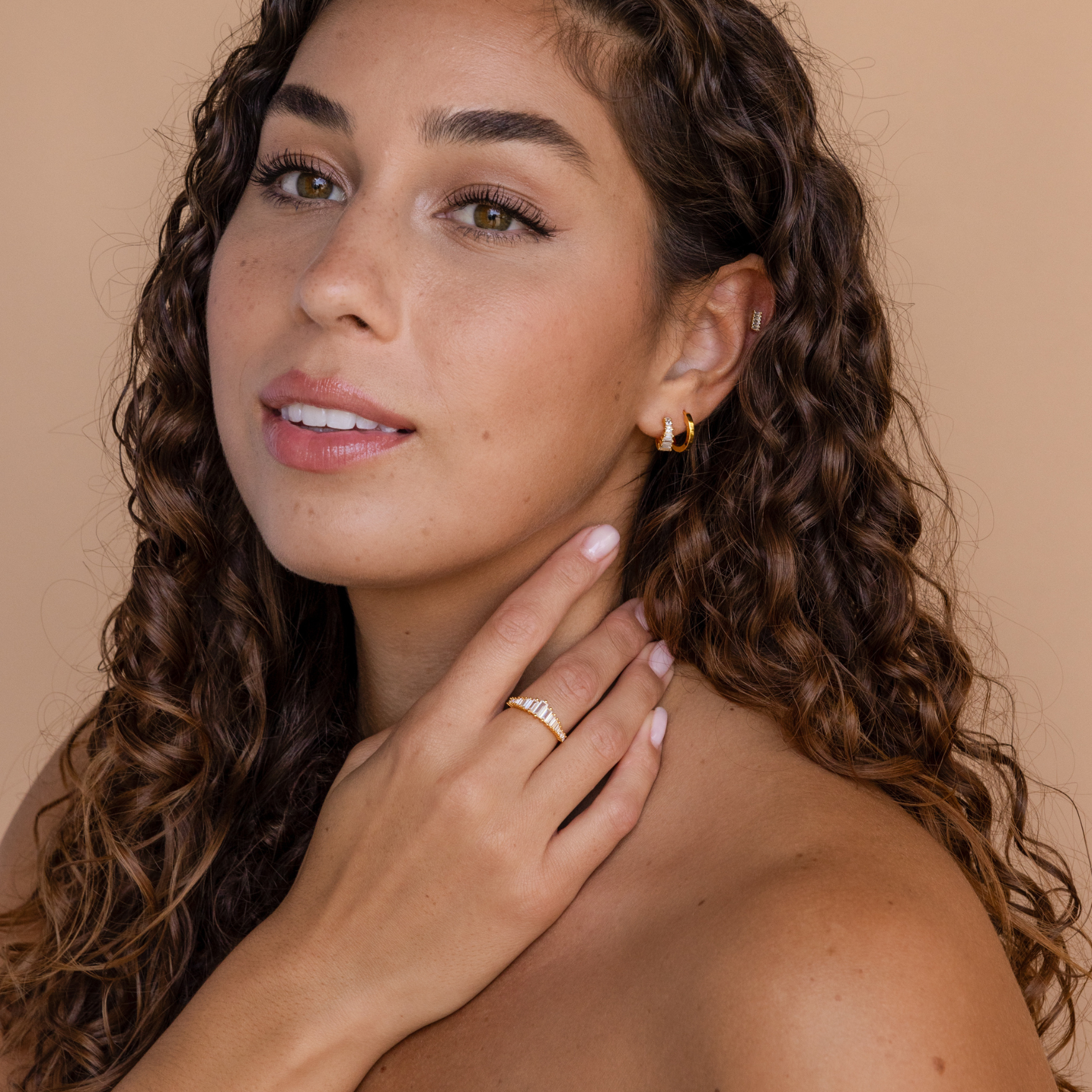 A woman with curly hair wears the Art Deco Huggies & Ring Set, posing with her hand near her neck against a beige background.