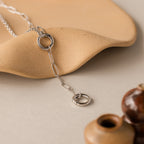 The Rolo Lariat Paperclip Clasp Necklace, featuring silver round links, is displayed on a beige textured surface beside small brown pottery.