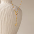 The Rolo Lariat Paperclip Clasp Necklace in 18K Gold features two interlocking rings on a paperclip chain, showcased in front of a white, textured ceramic vase.