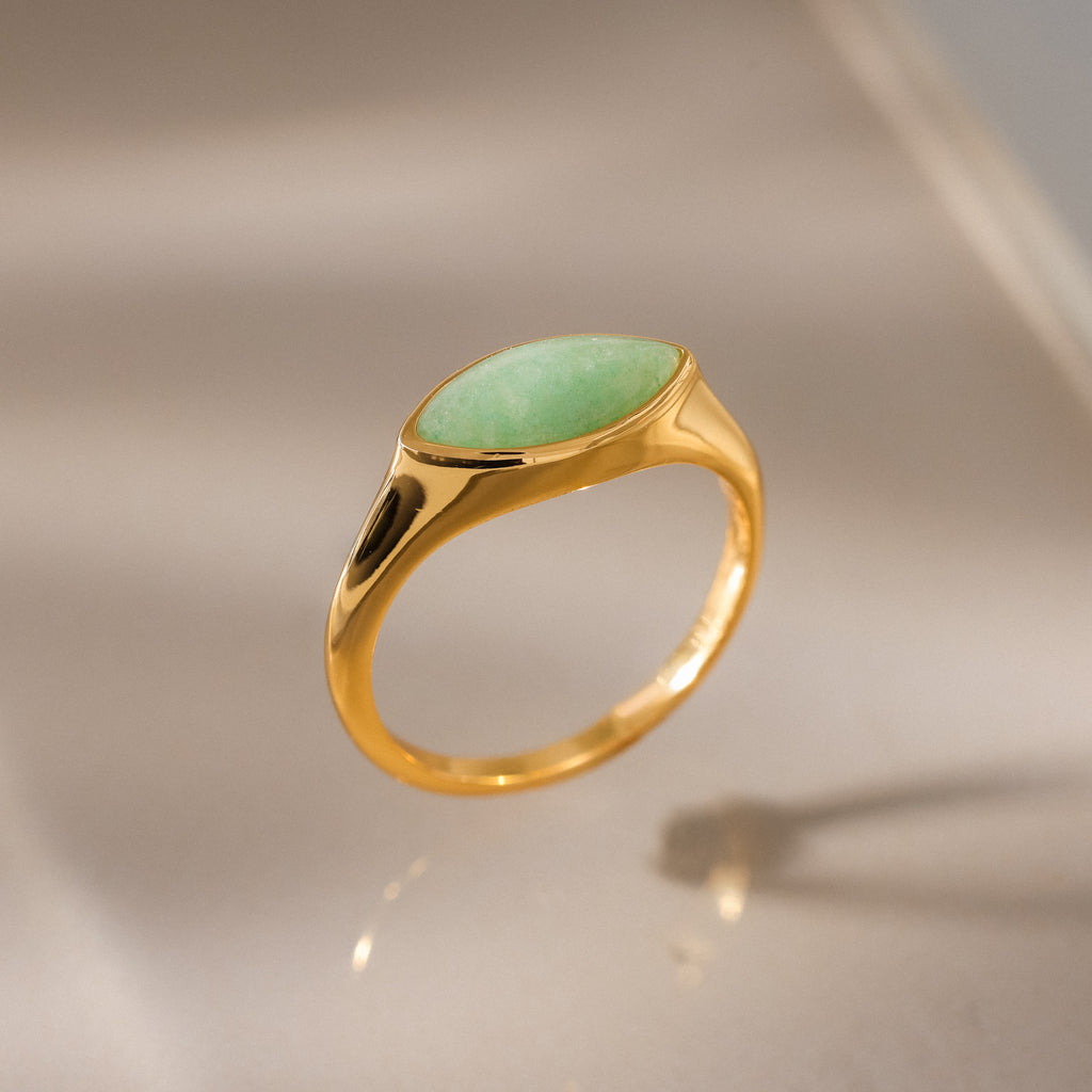 The Marquise Jade Signet Ring, featuring an oval green stone, is shown upright on a reflective beige surface with soft lighting.