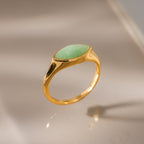 The Marquise Jade Signet Ring, featuring an oval green stone, is shown upright on a reflective beige surface with soft lighting.