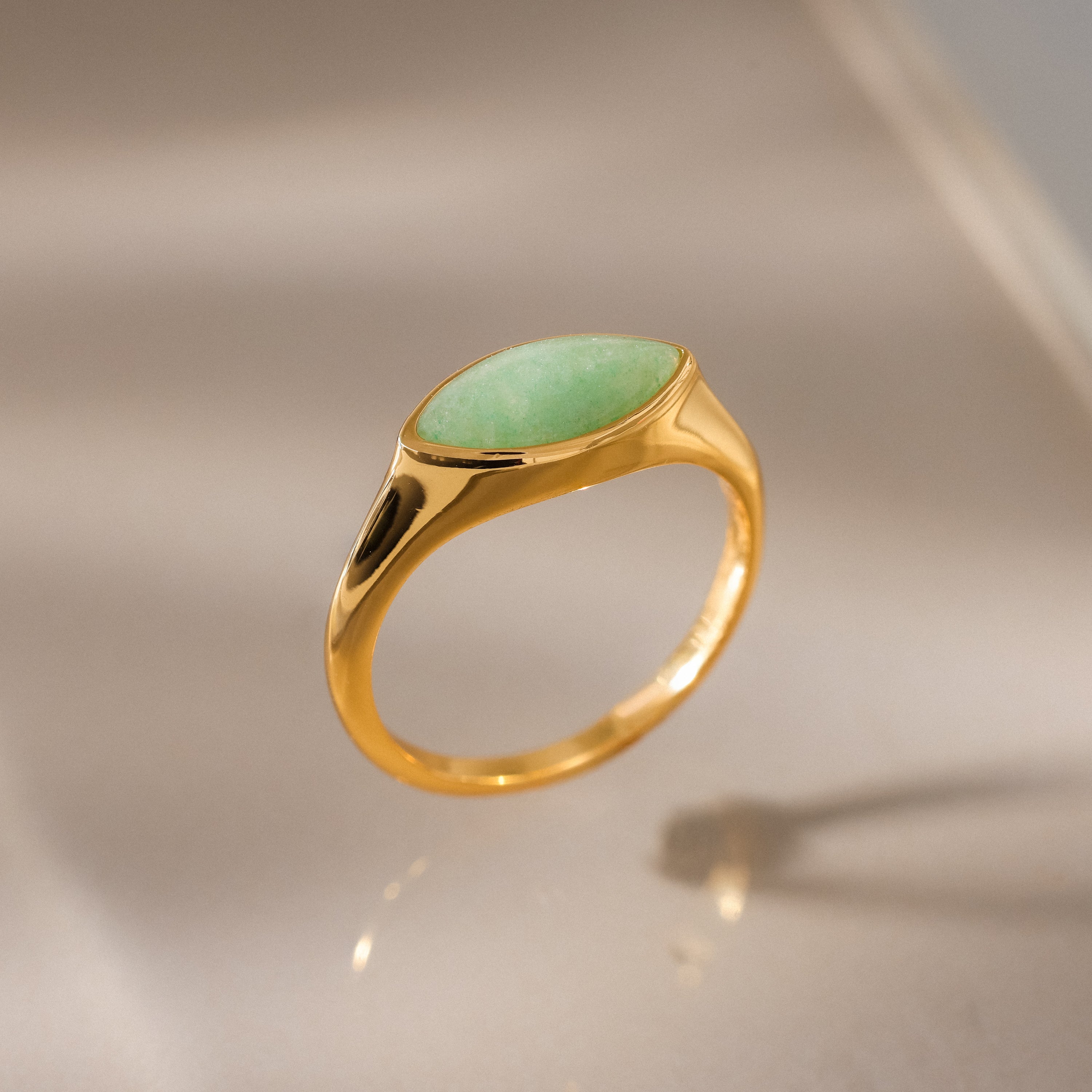 The Marquise Jade Signet Ring, featuring an oval green stone, is shown upright on a reflective beige surface with soft lighting.