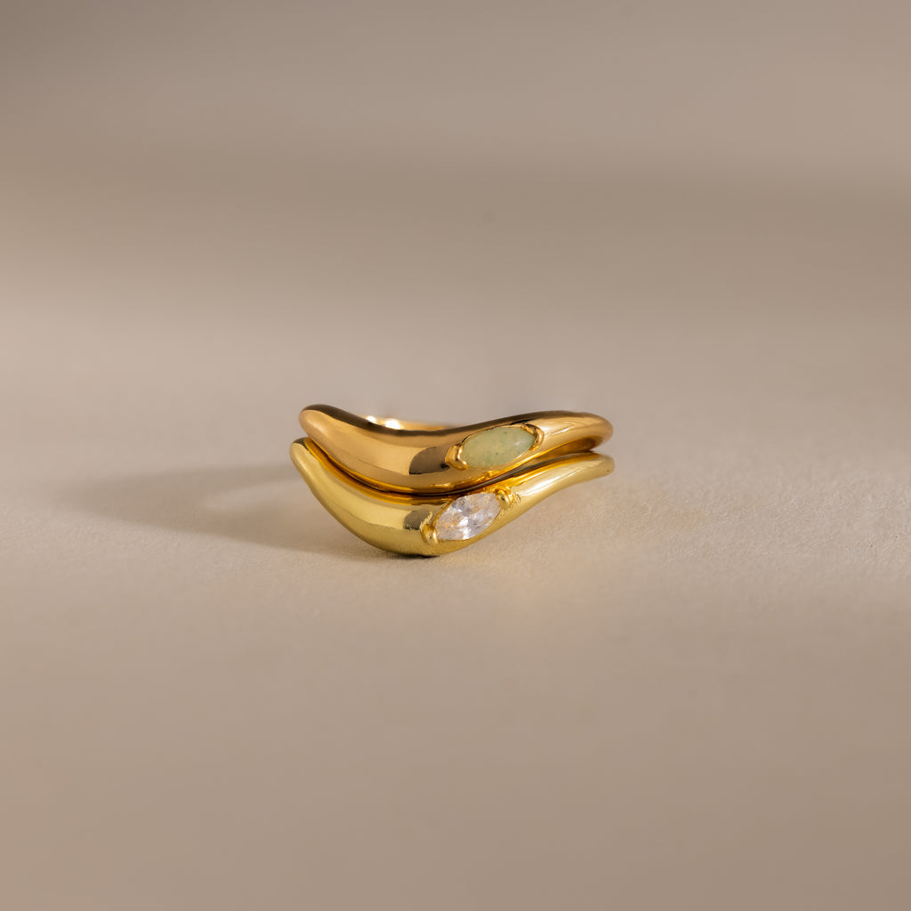 The Zen Gemstone Curve Ring, featuring a small diamond and minimalist gold band, is displayed alongside a light green gemstone-accented ring on a neutral background.