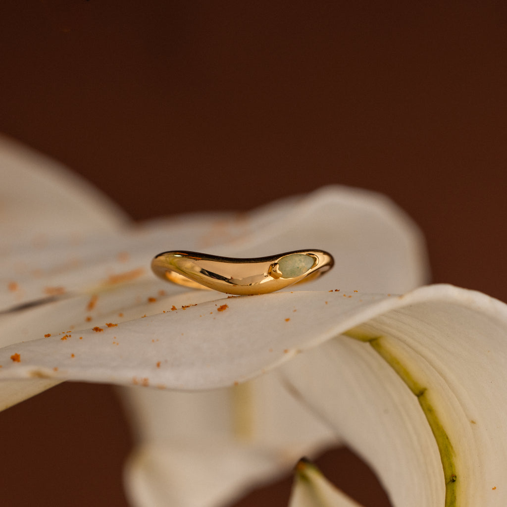 The Zen Jade Curve Ring in gold with a small stone is displayed on a white lily petal, set against a brown background.
