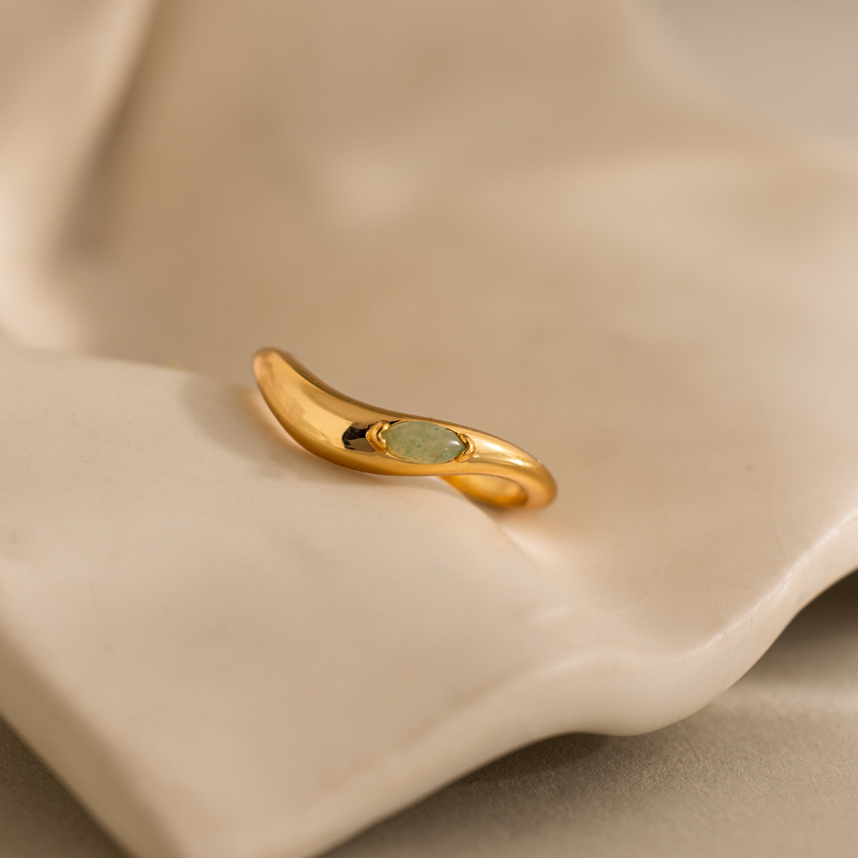 The Zen Gemstone Curve Ring, adorned with a delicate green gemstone accent, is displayed on a soft cream-colored fabric backdrop.