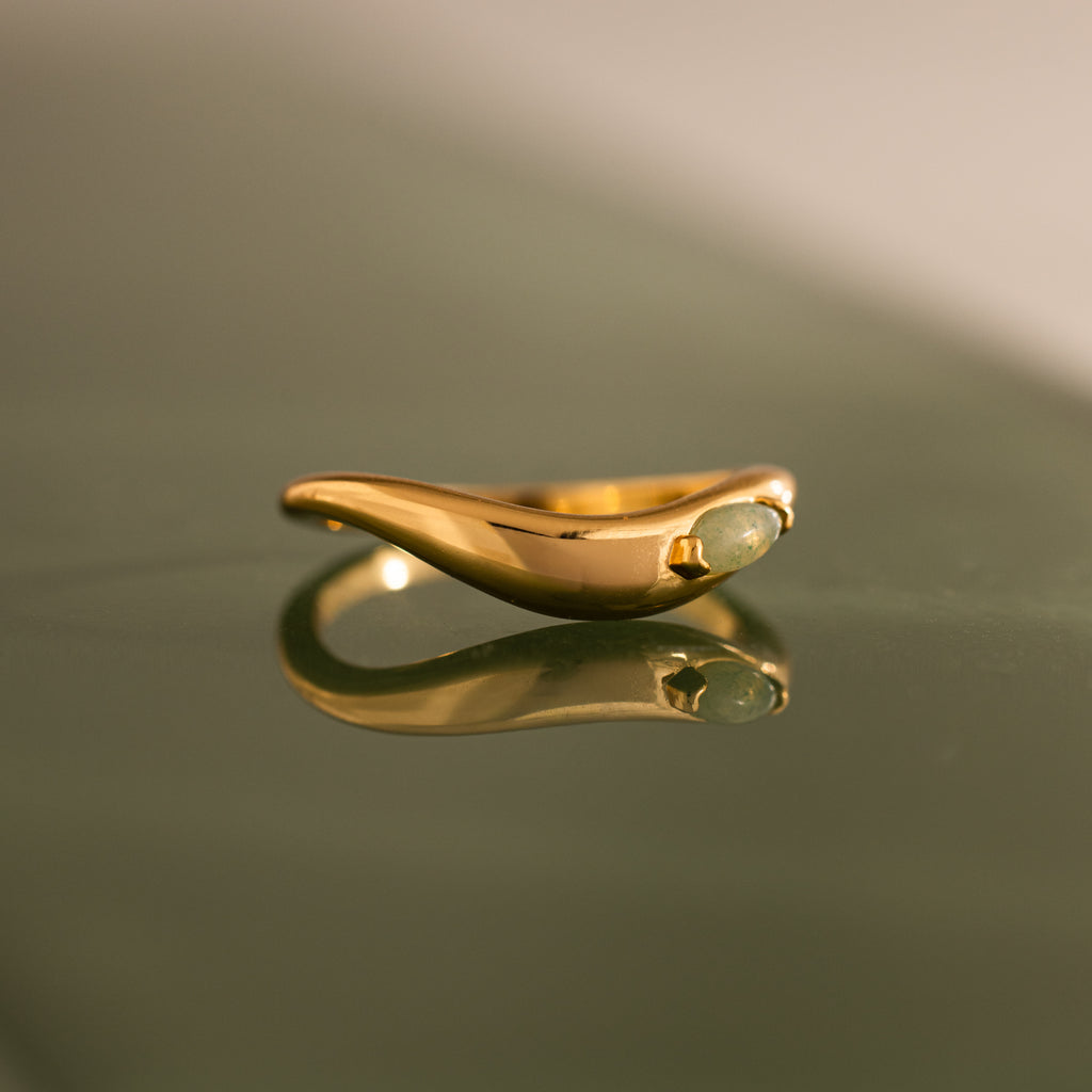 The Zen Jade Curve Ring showcases a smooth, sculpted band and a small green gemstone, elegantly reflected on glass.