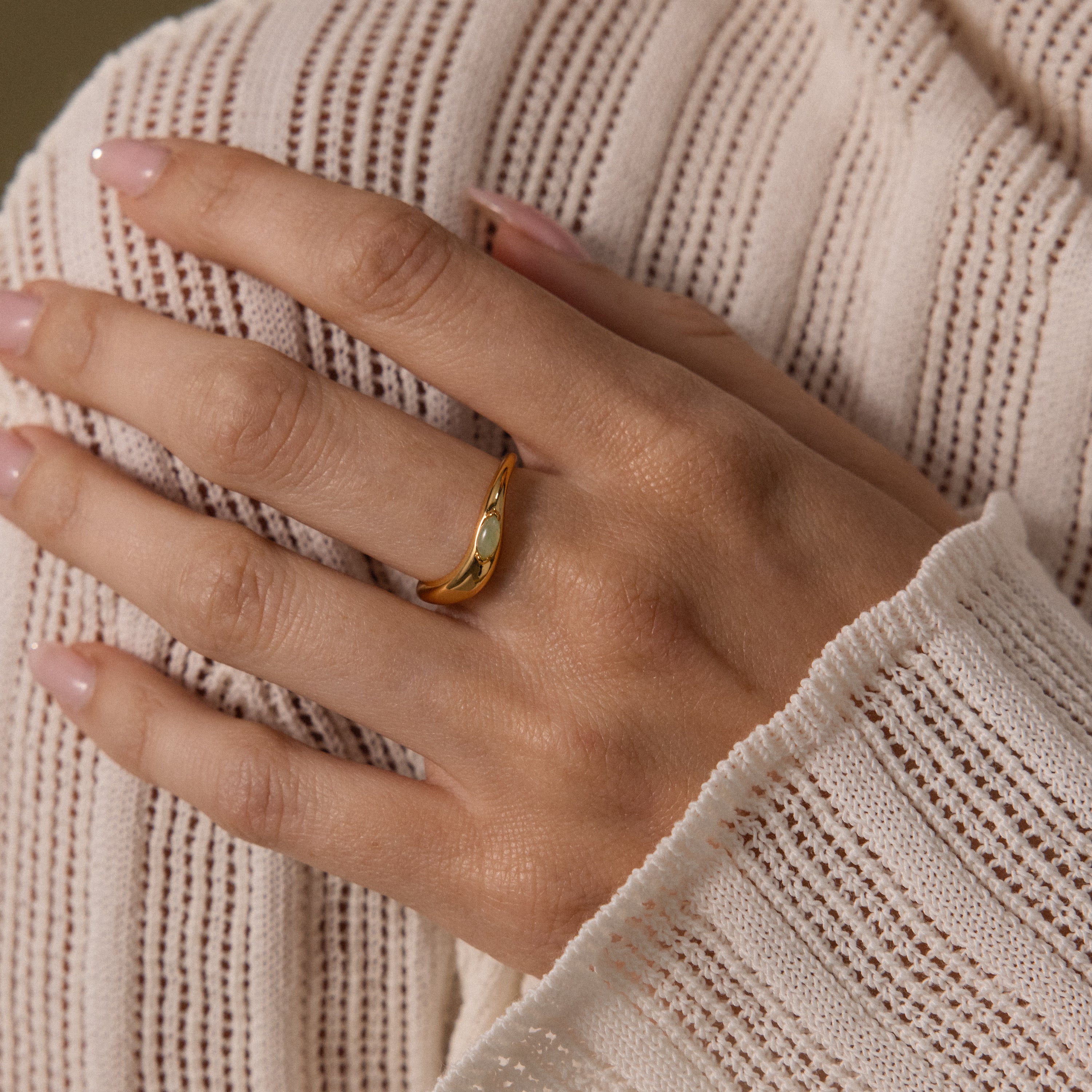 A hand with light pink nails wears the Zen Jade Curve Ring, its sculpted band and petite gem adding elegance against a cream knit sweater.