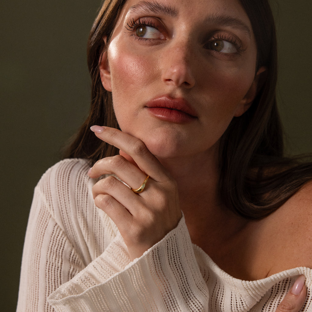 A woman with long brown hair and a white sweater gazes upward, her hand near her chin, elegantly displaying the Zen Jade Curve Ring on her finger.