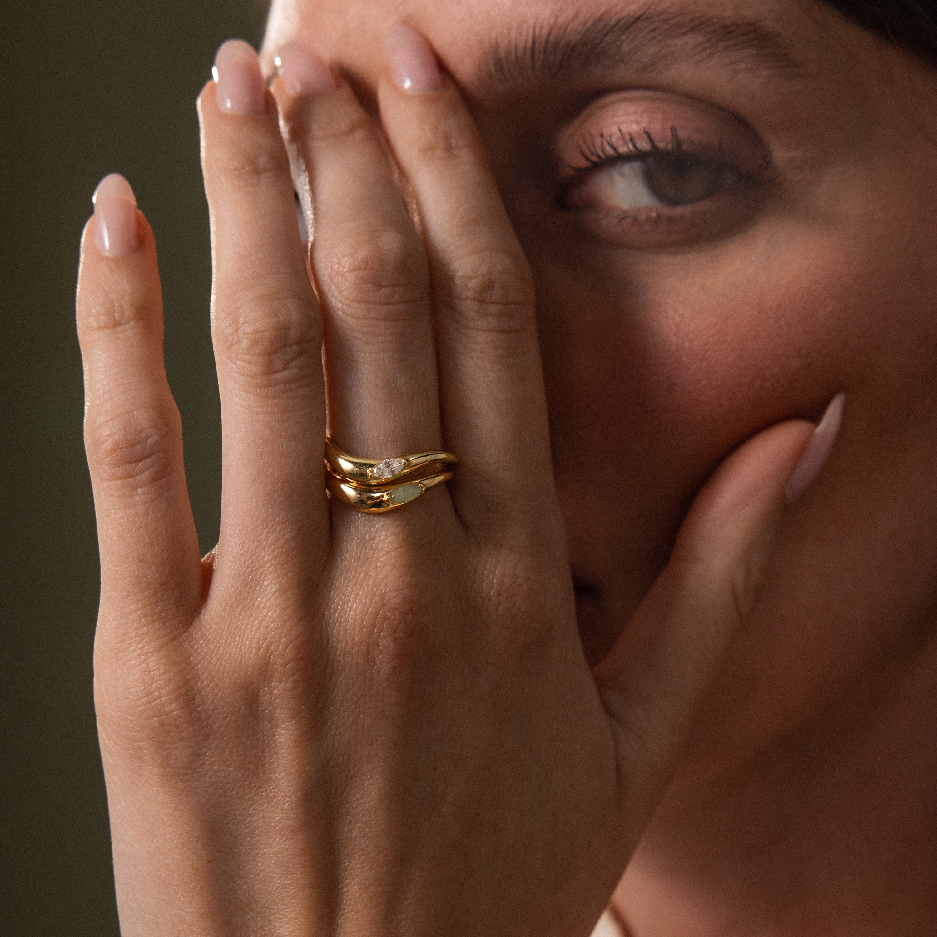 A woman with manicured nails covers half her face, displaying stacked gold rings—including the Zen Jade Curve Ring featuring a luminous jade gemstone—on her finger.