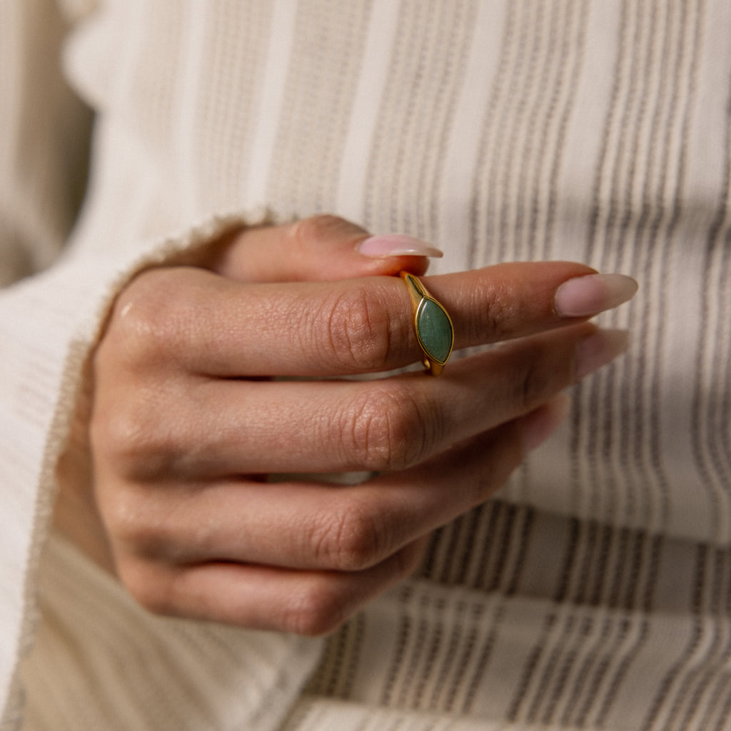 A hand with manicured nails wears the Marquise Jade Signet Ring, featuring a green stone, set against a striped fabric background.
