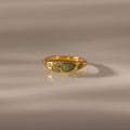 The Jade Dewdrop Ring features jade stones set in gold, radiating natural elegance as it rests gently on a smooth, light surface with soft shadows.