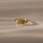 The Jade Dewdrop Ring features jade stones set in gold, radiating natural elegance as it rests gently on a smooth, light surface with soft shadows.