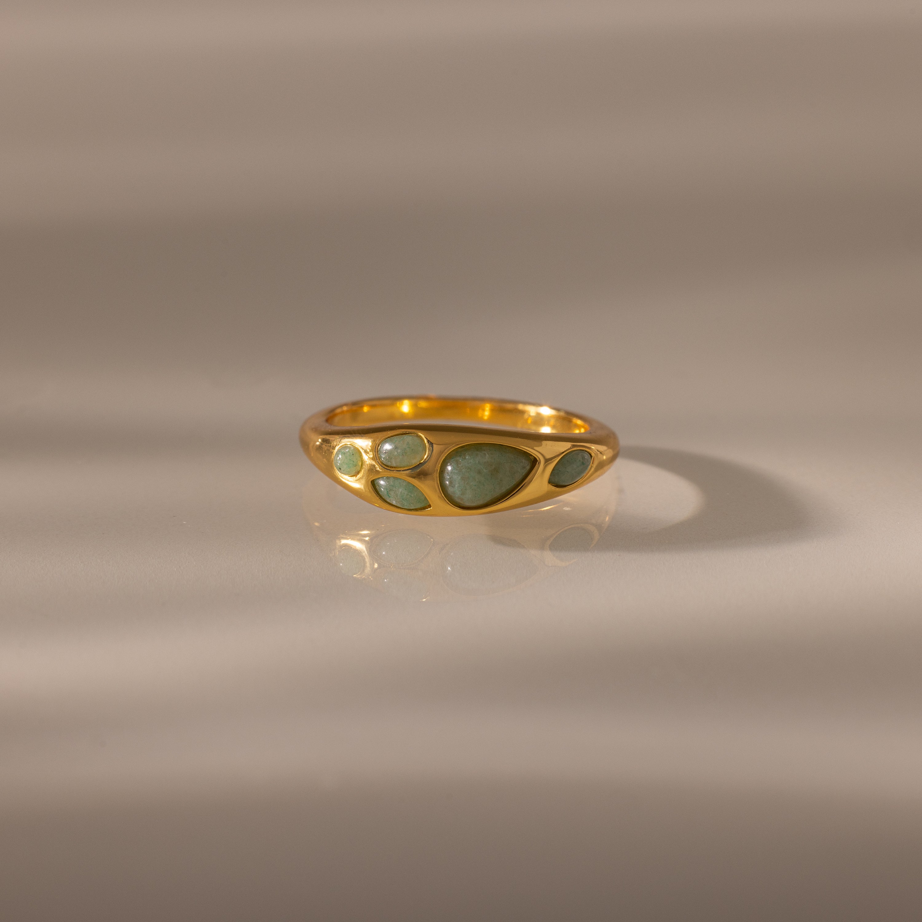 The Jade Dewdrop Ring features jade stones set in gold, radiating natural elegance as it rests gently on a smooth, light surface with soft shadows.
