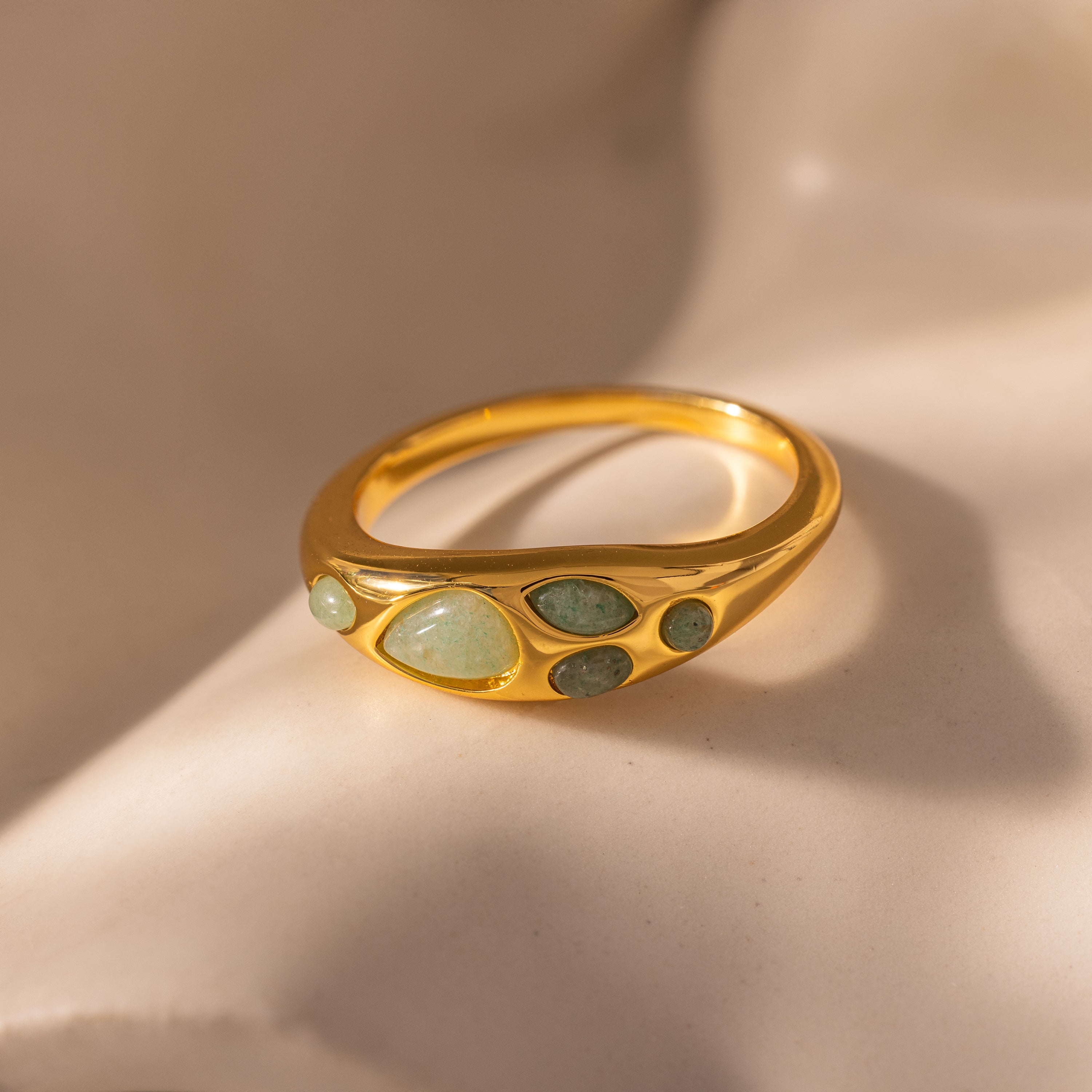 The Jade Dewdrop Ring showcases green gemstones in assorted shapes, highlighting natural elegance against a smooth beige background.