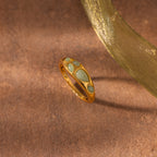 The Jade Dewdrop Ring, featuring green jade stone inlays, rests on a brown textured surface next to a sheer golden ribbon, highlighting its natural elegance.