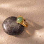 The Solace Jade Signet Ring, featuring an oval-cut jade stone, rests on a smooth black rock atop sandy ground.