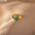 The Solace Jade Signet Ring features an oval-cut green stone set in gold, displayed on a textured beige surface with soft lighting—a modern heirloom.
