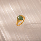 The Solace Jade Signet Ring, featuring an oval-cut green stone, rests on a light textured surface in soft lighting—a modern heirloom with timeless appeal.