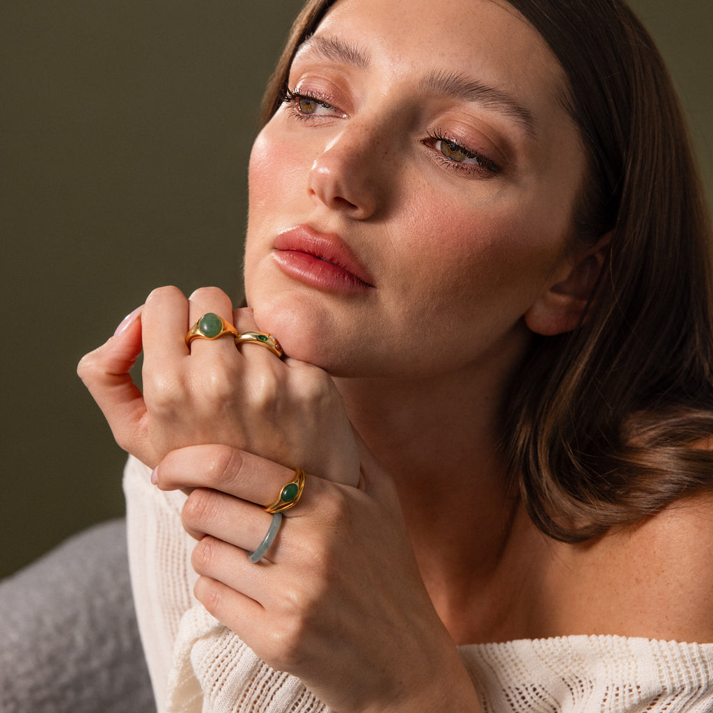 A woman with brown hair wears gold rings, including the Solace Jade Signet Ring, and a white off-shoulder top, gazing thoughtfully to the side.