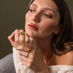 A woman with brown hair wears gold rings, including the Solace Jade Signet Ring, and a white off-shoulder top, gazing thoughtfully to the side.