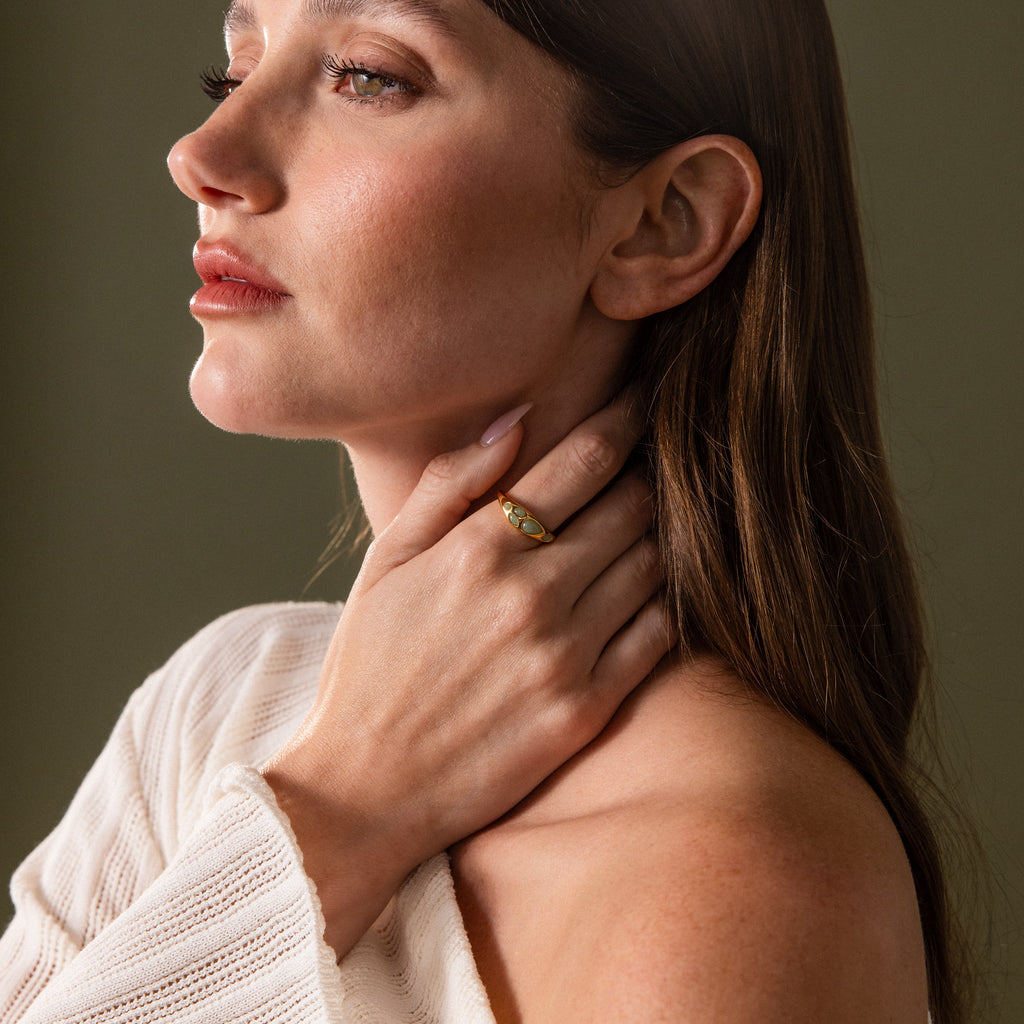 A woman in a white sweater touches her neck, highlighting natural elegance with the Jade Dewdrop Ring featuring subtle jade stones, set against a dark background.