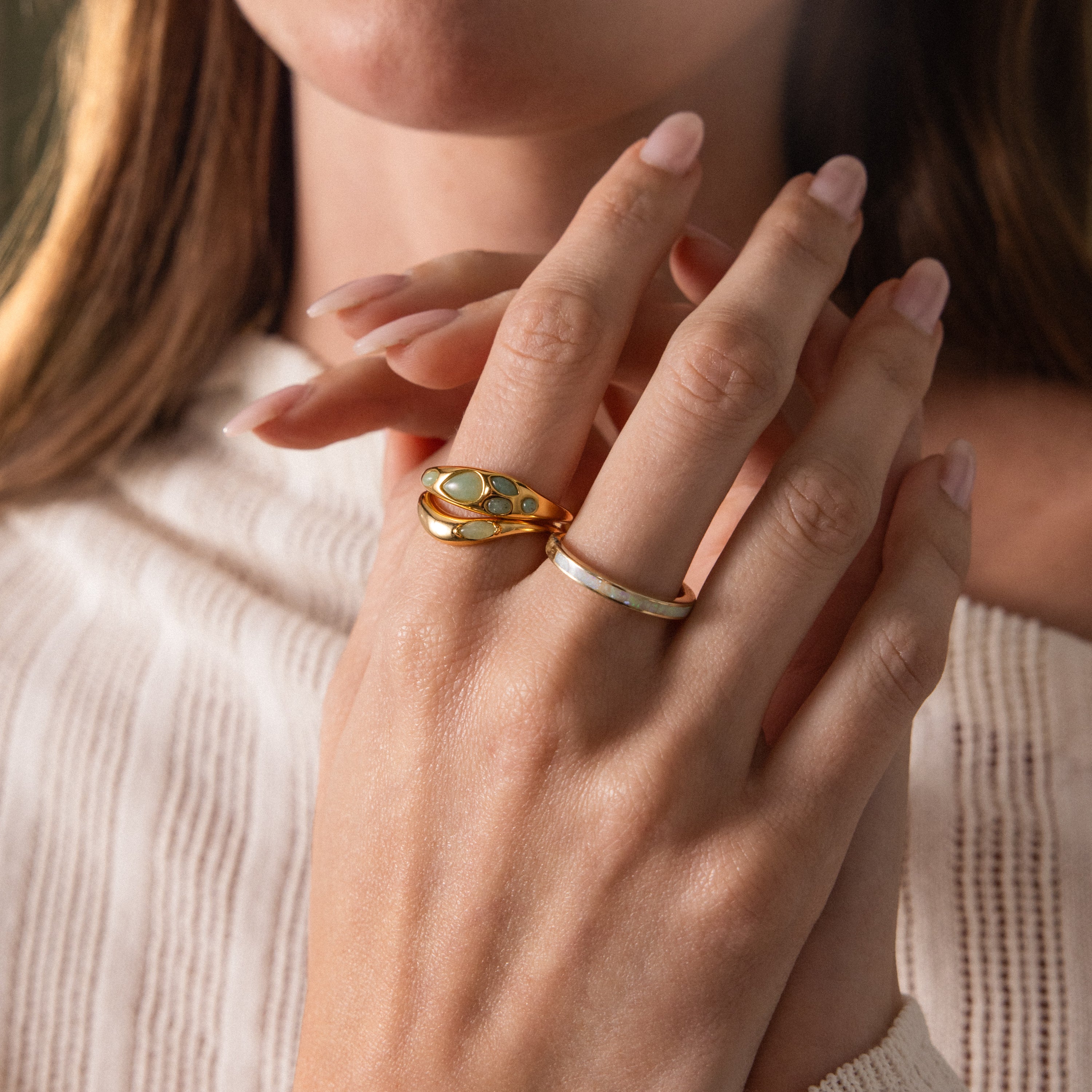 A womans hand with natural nails displays elegance, wearing two gold rings—one being the Jade Dewdrop Ring—held gently near her face.