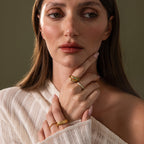 A woman with long brown hair wears gold rings and the Jade Dewdrop Ring, her white textured top highlighting her elegance as she gazes thoughtfully to the side.