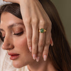 A woman with long brown hair rests her hand on her face, wearing the Solace Jade Signet Ring, a modern heirloom gold ring featuring an oval-cut jade stone.