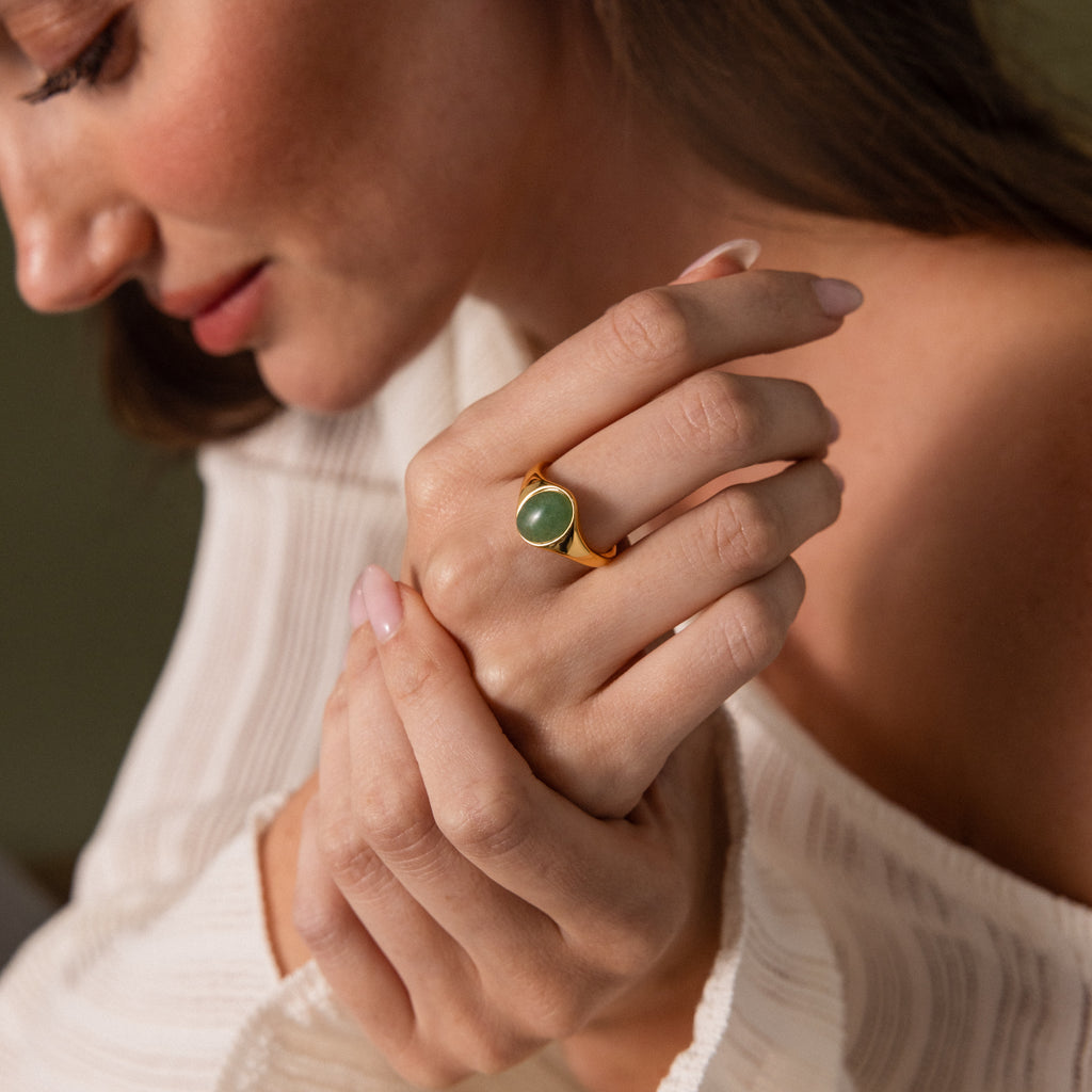 A woman in a white top gently touches her hand while wearing the Solace Jade Signet Ring, featuring a gold band and green stone—a true modern heirloom.