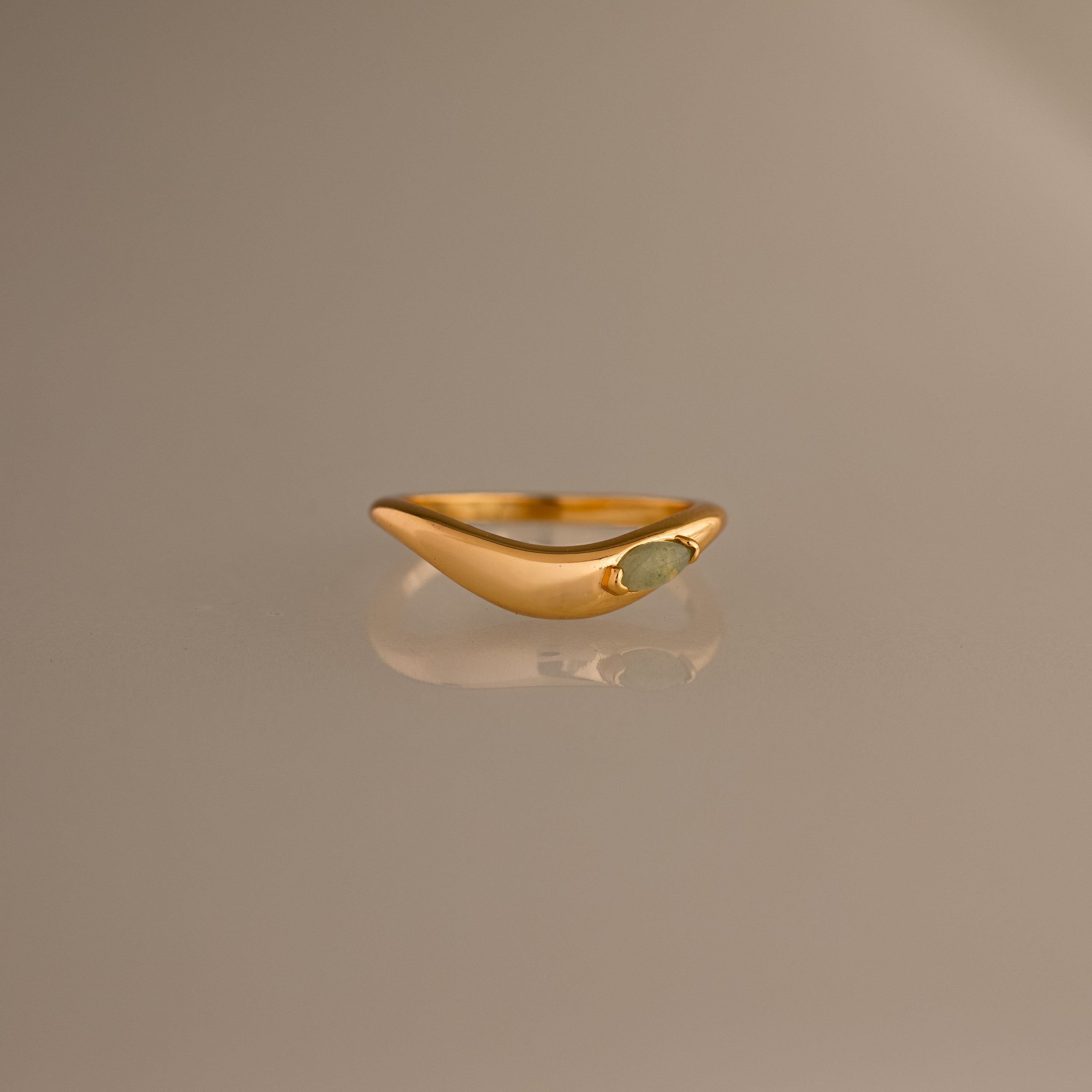 The Zen Jade Curve Ring features a gold sculpted band with a smooth, curved design and a small oval stone set on one side, shown against a neutral background.