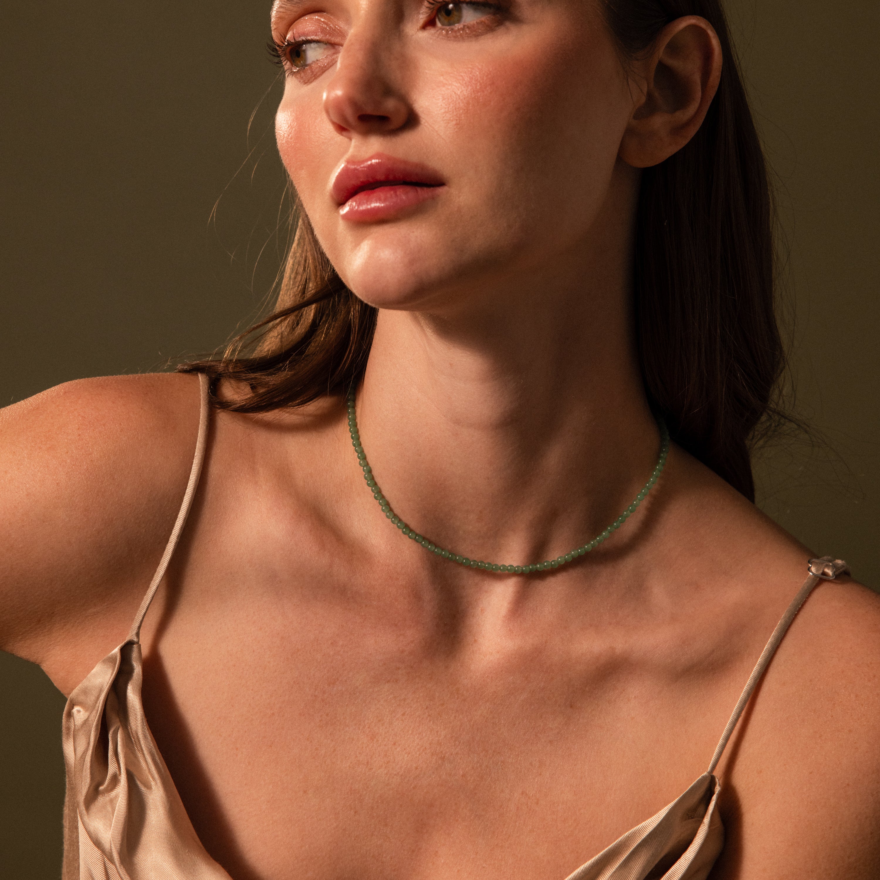 A woman in a satin dress wears the Tiny Jade Choker Necklace in 18K Gold, gazing to the side against a neutral backdrop.