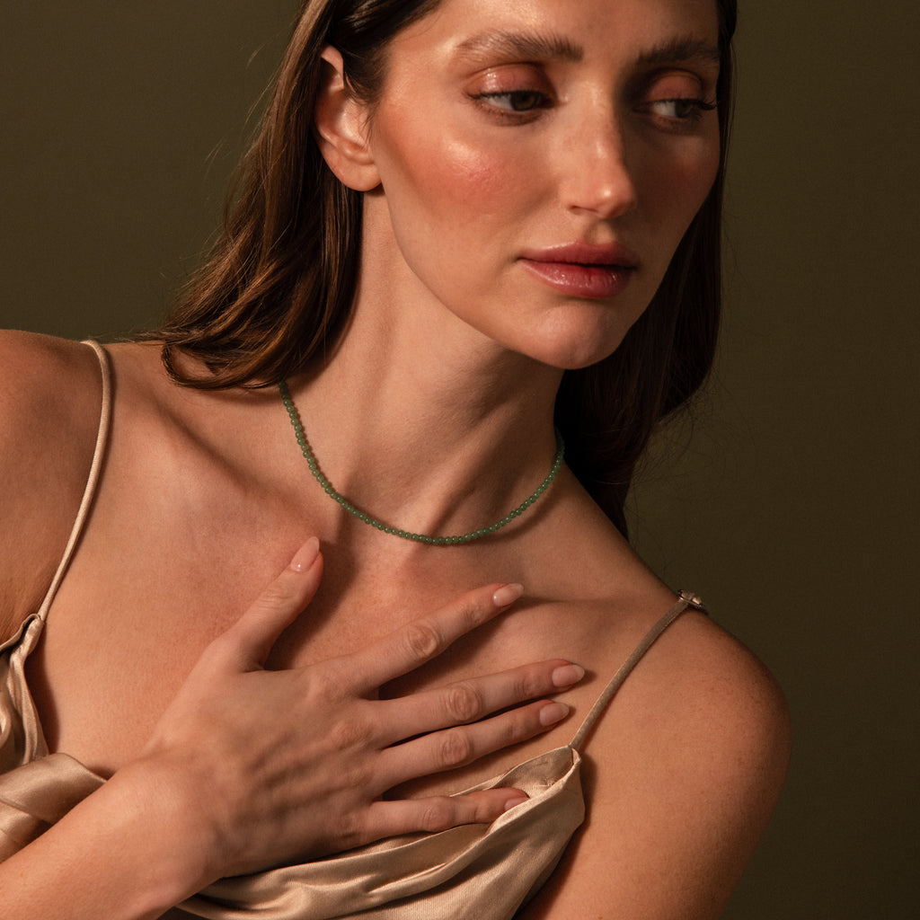 A woman in a satin dress touches her chest, highlighting the Tiny Jade Choker Necklace against an olive background.