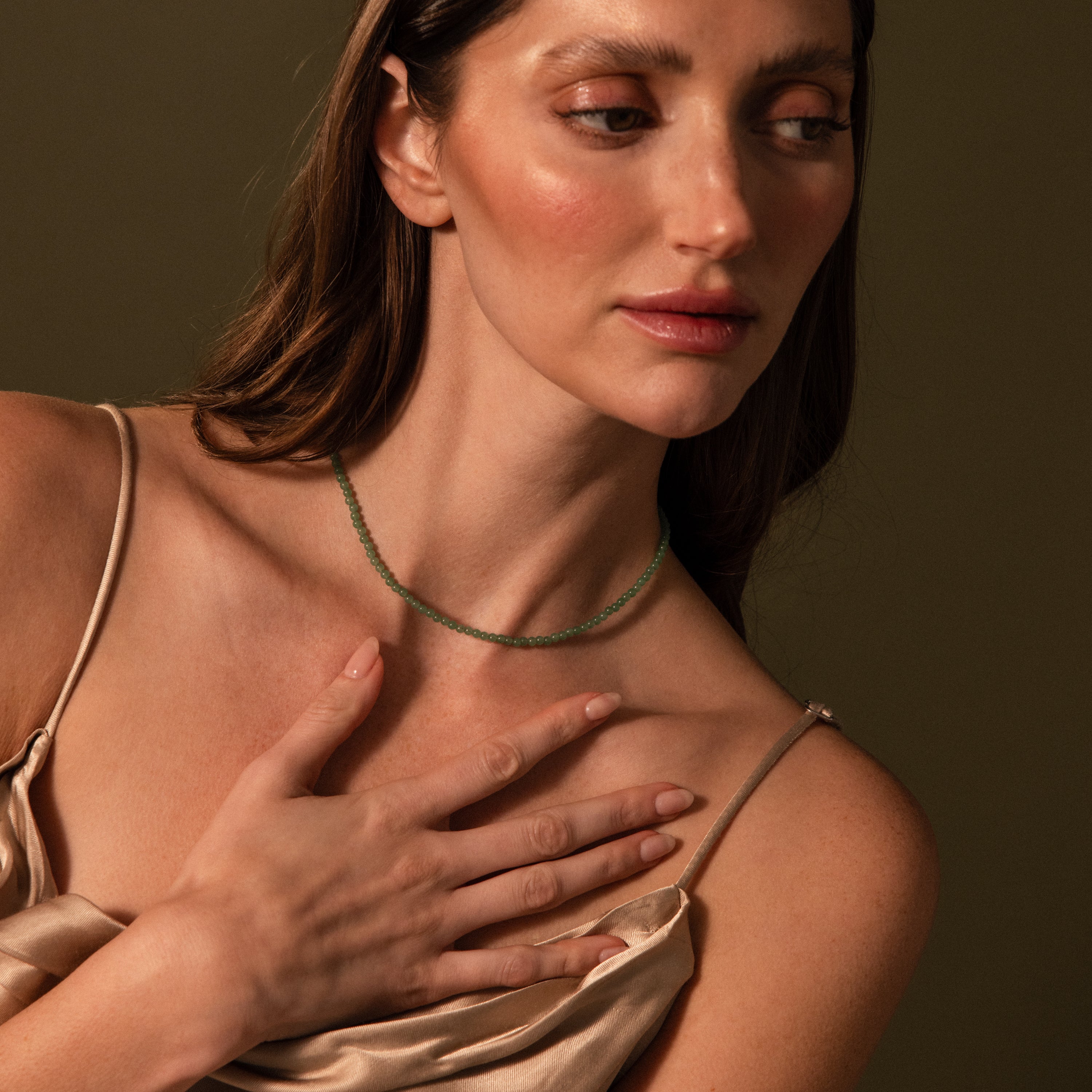 A woman in a satin dress touches her chest, highlighting the Tiny Jade Choker Necklace against an olive background.