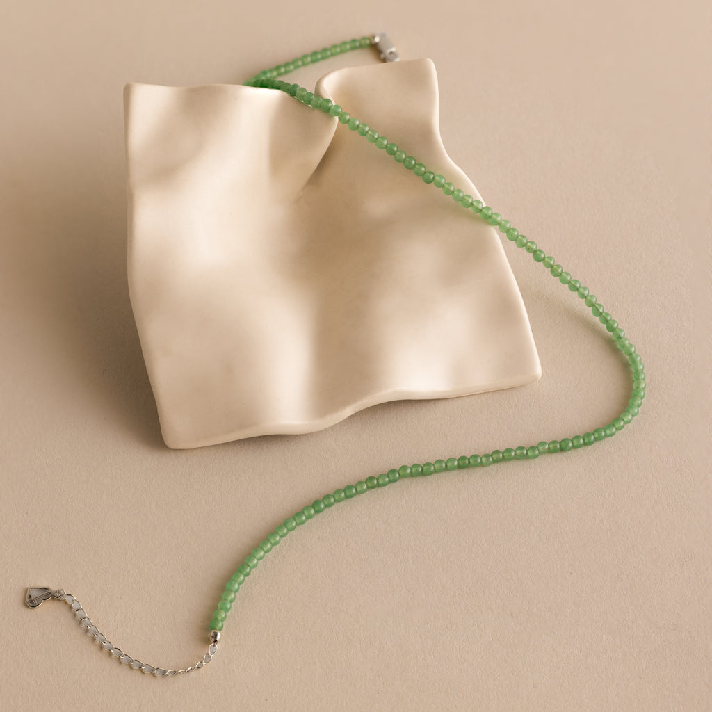 The Tiny Jade Choker Necklace, featuring green beads and a layered design, is displayed on an abstract wavy cream ceramic dish.
