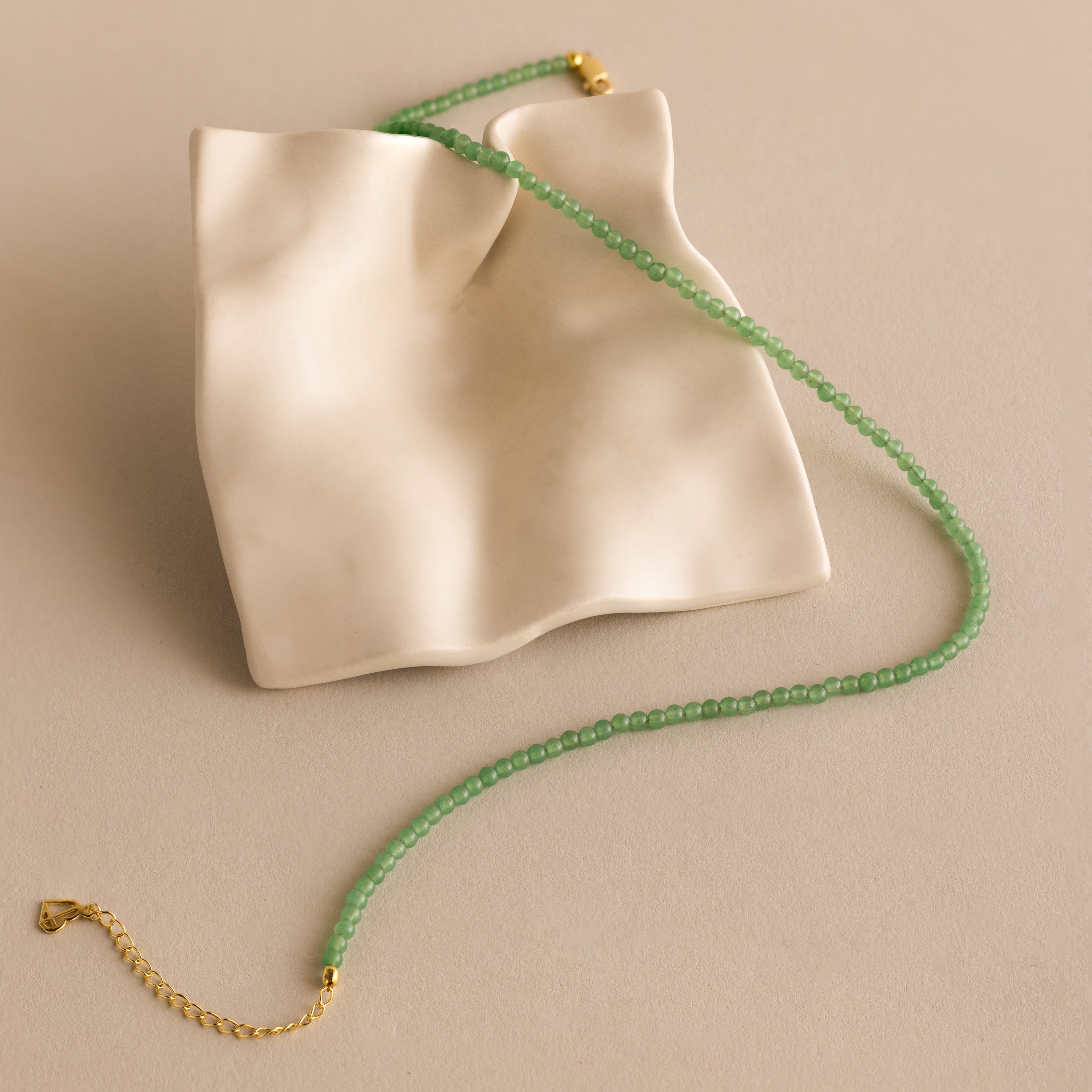 The Tiny Jade Choker Necklace in 18K Gold, with green jade beads and a gold clasp, is displayed on a wavy cream-colored ceramic tray.