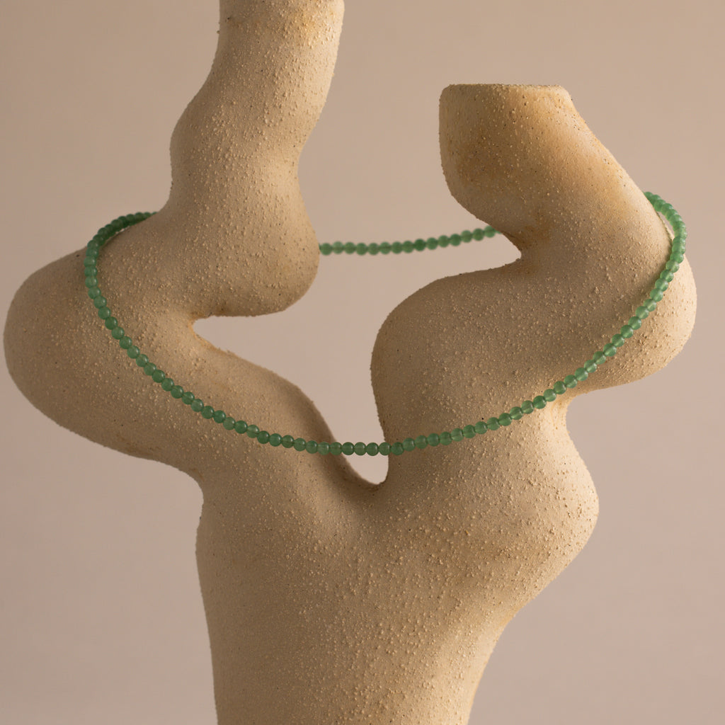 The Tiny Jade Choker Necklace, featuring green beads in layered strands, is draped over an abstract beige stone sculpture set against a plain background.