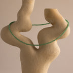 The Tiny Jade Choker Necklace, featuring green beads in layered strands, is draped over an abstract beige stone sculpture set against a plain background.