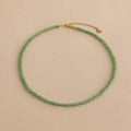 The Tiny Jade Choker Necklace in 18K Gold features delicate green jade beads and a gold clasp with extender, beautifully presented against a beige backdrop.