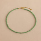 The Tiny Jade Choker Necklace in 18K Gold features delicate green jade beads and a gold clasp with extender, beautifully presented against a beige backdrop.