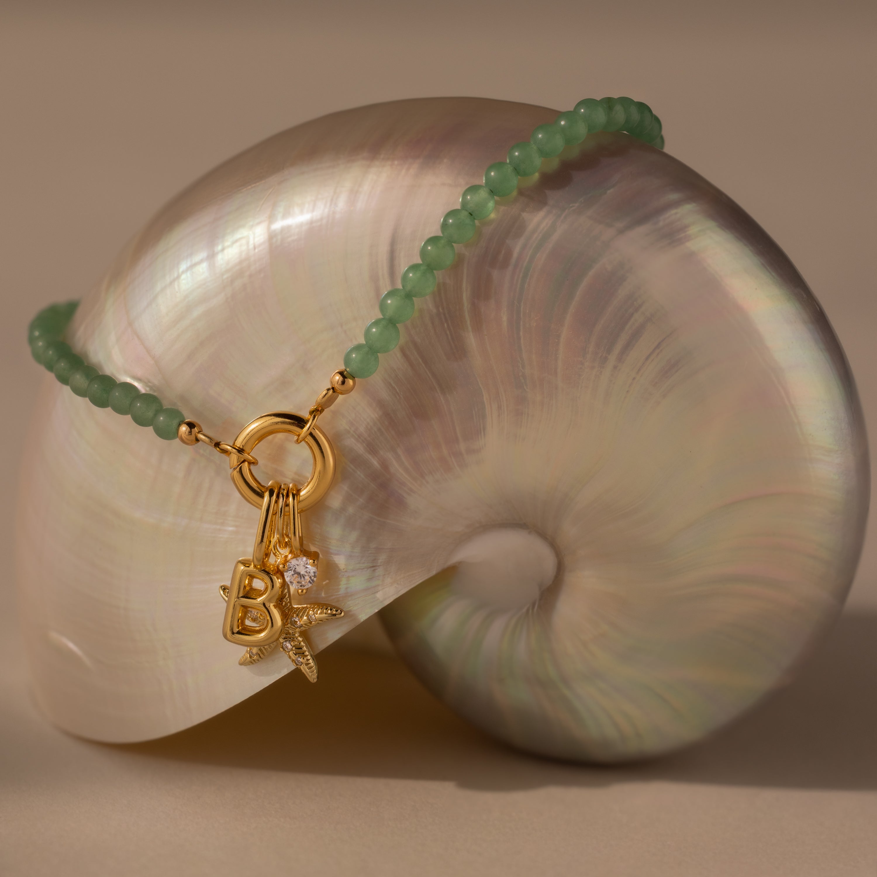 The Beaded Jade Clasp Bracelet, featuring green beads and gold charms, is displayed on a shiny spiral seashell background.