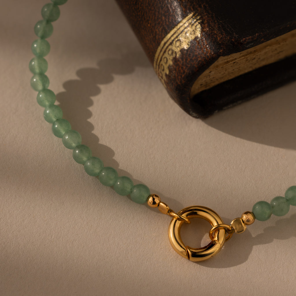 The Beaded Jade Clasp Bracelet, featuring green jade accents and a gold clasp, rests beside the corner of a closed brown book adorned with gold detailing.